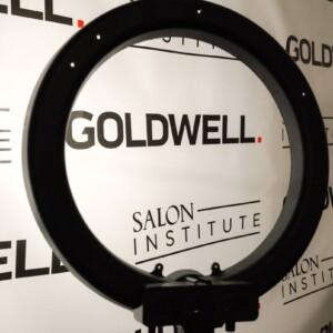A ring light stands in front of a backdrop featuring the Goldwell and Salon Institute logos.
