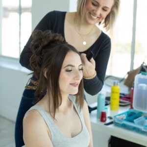 A woman styles another woman's hair in a bright room with beauty products on the table nearby.