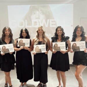 Five women in black dresses stand indoors, holding certificates and smiling in front of a presentation screen that reads "GOLDWELL.