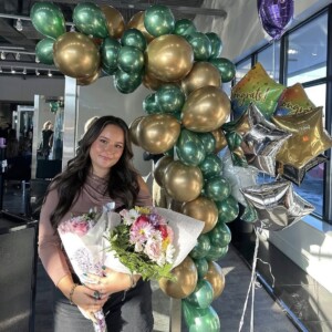 Screenshot A woman holding a bouquet of flowers stands in front of green and gold balloons and "Congrats" balloons.