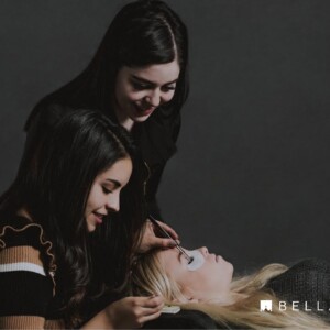 Two women apply eyelash extensions to a woman lying down, with Bella Lash branding visible in the corner.