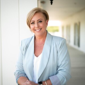 Woman in a light blue blazer stands on a porch, smiling at the camera, with a blurred outdoor background.