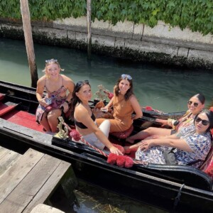 Five women sit and smile in a gondola on a canal, with greenery and a stone wall in the background.