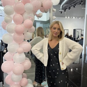 Screenshot A woman in a black dress and white cardigan stands beside a pink and white balloon display in a modern salon.