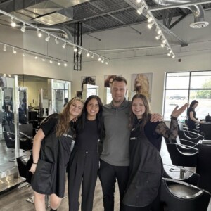 Four people pose together and smile inside a modern hair salon with black chairs and bright overhead lighting.
