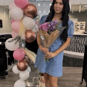 Screenshot Woman in a denim dress holding a bouquet of flowers stands in front of a pink and white balloon arch indoors.