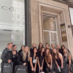 A group of people in black outfits stand outside a building entrance, holding branded tote bags and smiling at the camera.