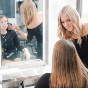 A hairstylist styles a client's hair in a salon, both facing a large mirror with bright natural light.
