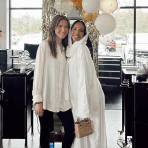 Two women standing and smiling indoors, with balloons and gold streamers in the background.