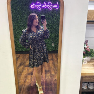 Woman takes a mirror selfie wearing a floral dress and wedge sandals in a room with a neon sign and greenery wall.