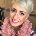 Woman with short blonde hair, bright pink lipstick, and a pink textured scarf smiles at the camera indoors.