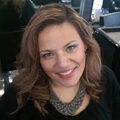 Woman with wavy light brown hair, wearing a dark top and statement necklace, smiling at the camera indoors.
