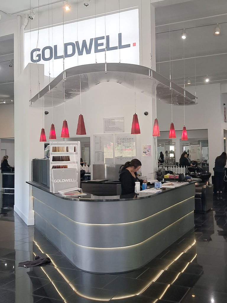 Reception desk at a modern hair salon with red pendant lights and a large "GOLDWELL" sign overhead.