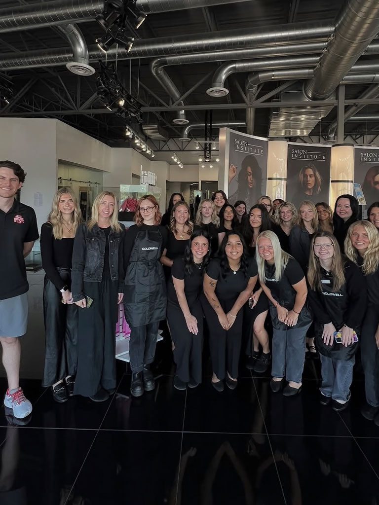 A group of people in black uniforms pose together inside a modern salon with posters and styling stations in the background.