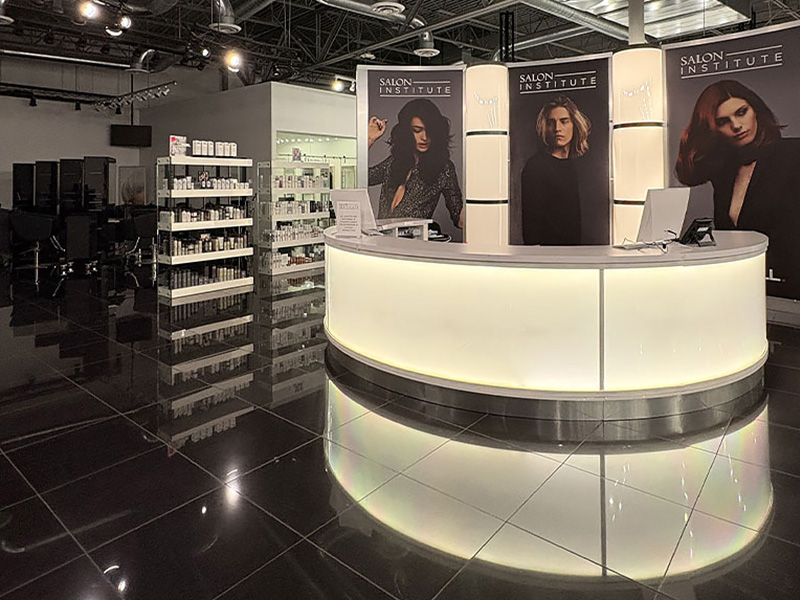 Modern salon reception area with a curved lighted desk, hair product shelves, and posters of women on the wall.