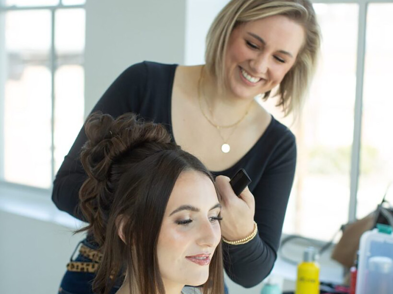A hairstylist is styling a woman's hair in a bright, modern salon with beauty products visible on the counter.