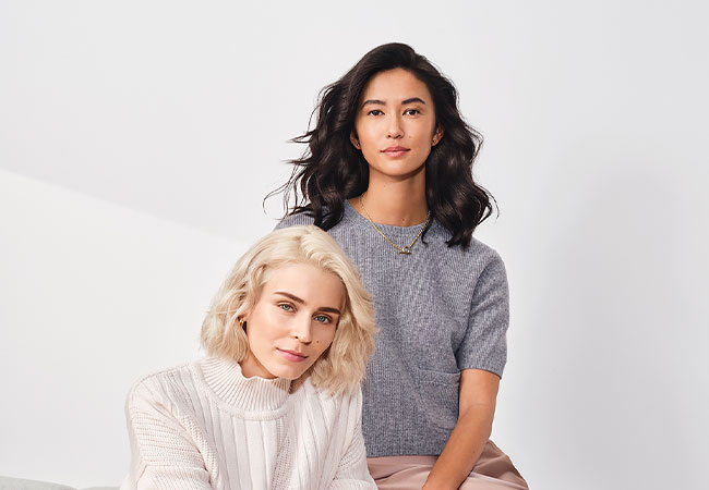 Two women pose together, one seated with blonde hair and a white sweater, the other standing with dark hair and a gray top.