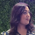 Woman with wavy brown hair and floral top standing in front of a green leafy background, looking to the right.