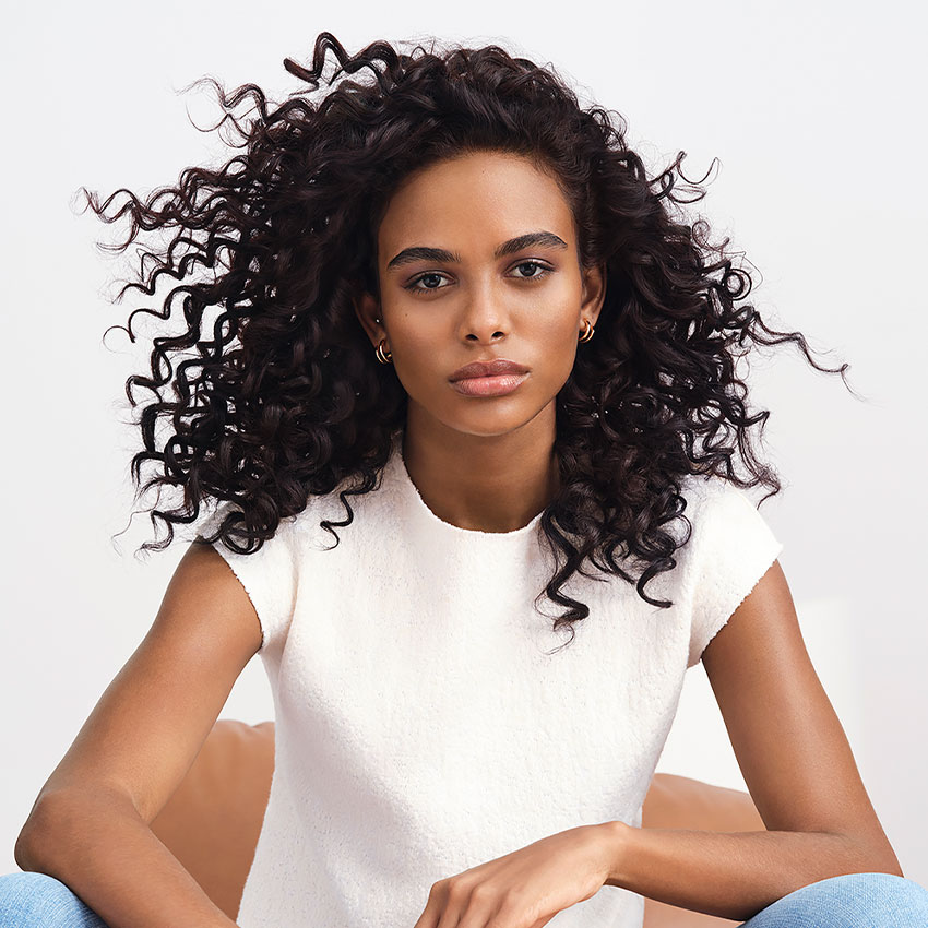 Woman with long, curly dark hair wearing a short-sleeve white top, sitting and looking directly at the camera.