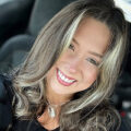 Woman with long, wavy hair and light highlights smiles inside a car, wearing a black top and a necklace.